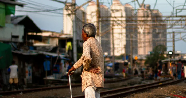 seorang pria tua berdiri di dekat rel kereta api dengan latar rumah-rumah gubuk