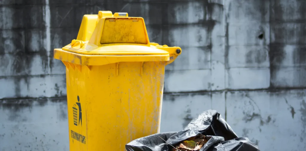 tempat sampah berwarna kuning dengan kantong plastik berisi sampah di sebelahnya