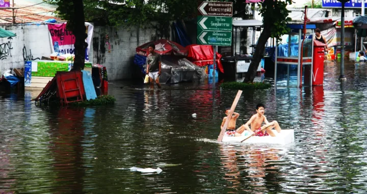 dua anak lelaki di perahu kayu kecil sedang mendayung di tengah jalanan penuh air banjir