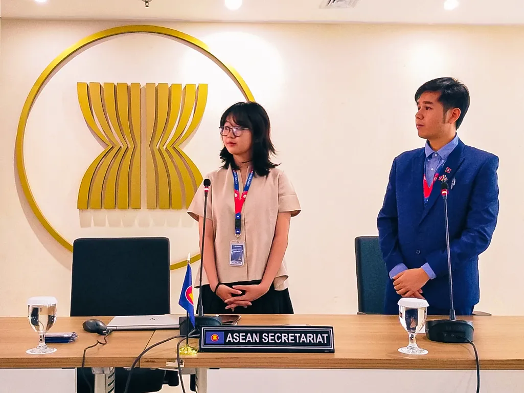 seorang perempuan dan seorang laki-laki muda berdiri dan melakukan presentasi di depan logo ASEAN.