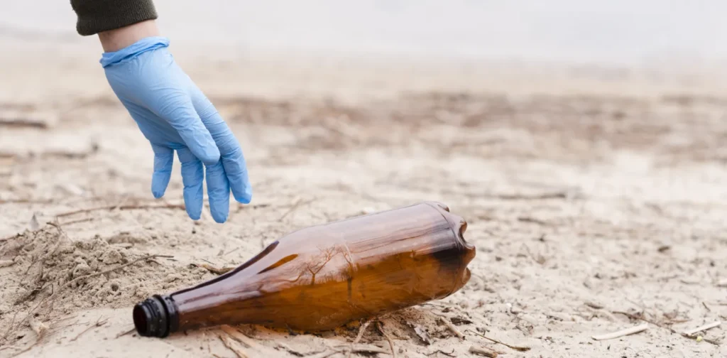 tangan dengan sarung tangan berwarna biru mengambil botol plastik berwarna coklat di atas pasir