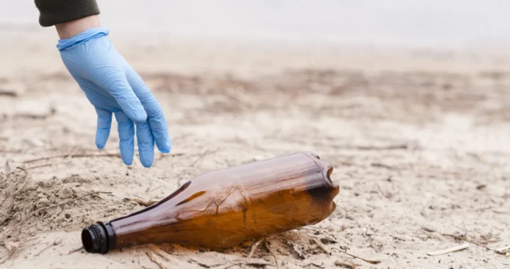 tangan dengan sarung tangan berwarna biru mengambil botol plastik berwarna coklat di atas pasir