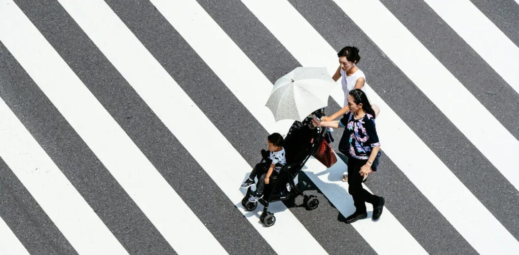 dua perempuan dan seorang anak di stroller berjalan di zebra-cross jalan dengan salah seorang memegang payung