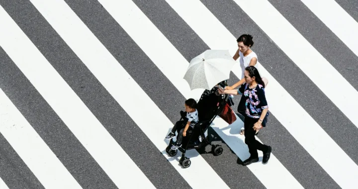 dua perempuan dan seorang anak di stroller berjalan di zebra-cross jalan dengan salah seorang memegang payung