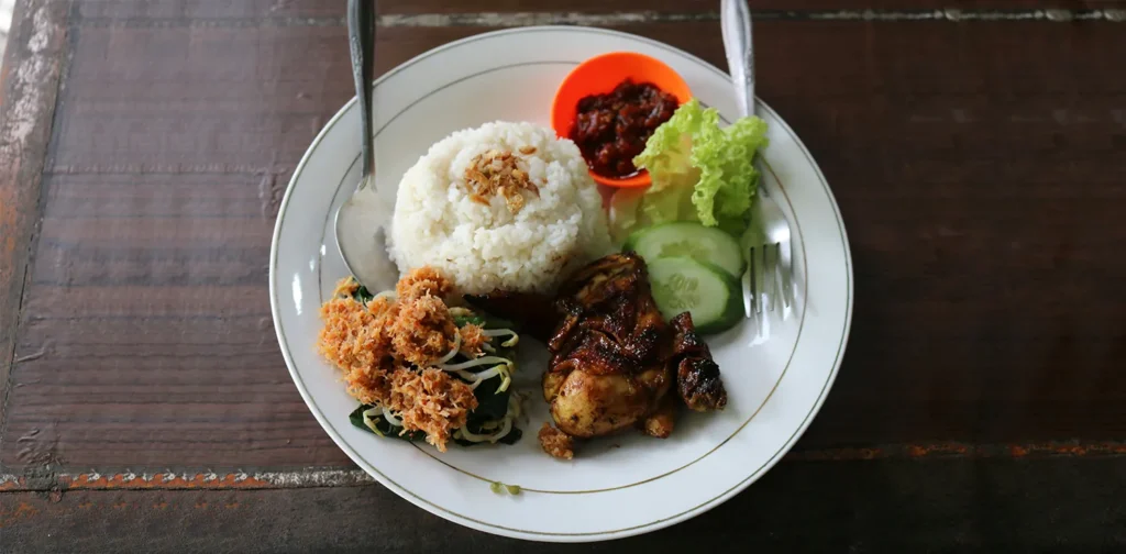 piring makan berisi nasi dengan lauk ayam, urap, sambal, dan irisan timun dan selada