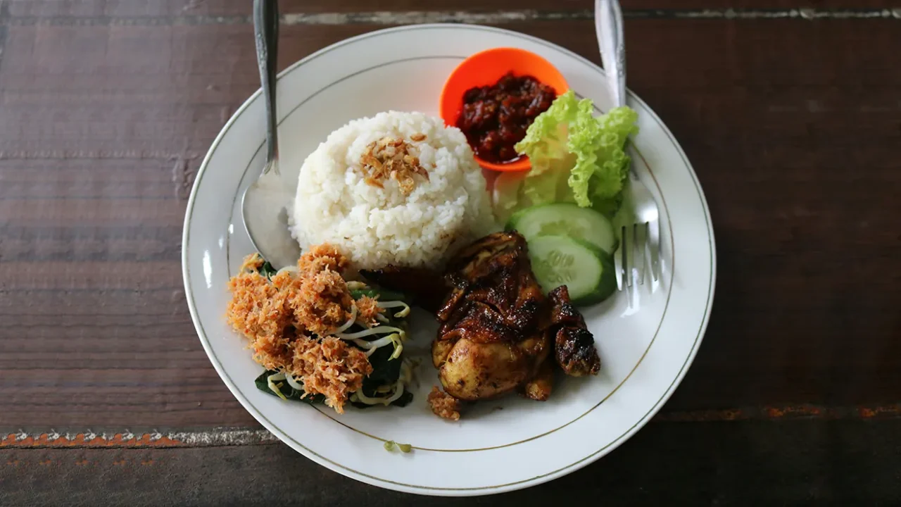 piring makan berisi nasi dengan lauk ayam, urap, sambal, dan irisan timun dan selada