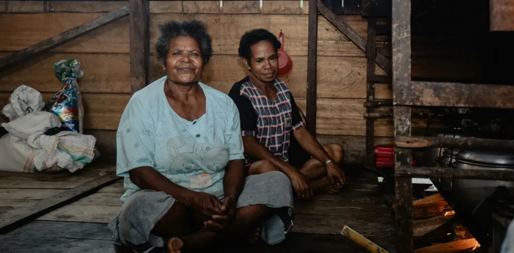 dua perempuan Papua duduk di dapur tradisional dengan lantai dan tembok kayu dan tungku api