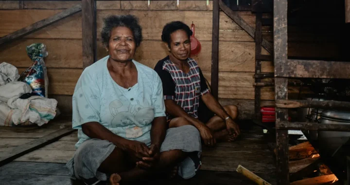 dua perempuan Papua duduk di dapur tradisional dengan lantai dan tembok kayu dan tungku api