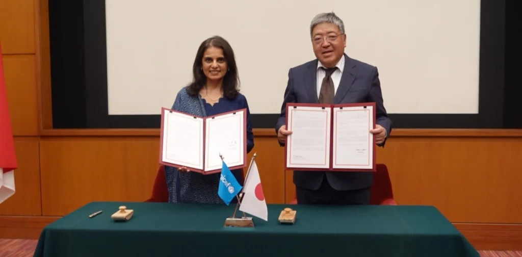 seorang perempuan dan seorang laki-laki menunjukkan dokumen yang dibentangkan dengan bendera kecil Jepang dan UNICEF di atas meja berkain hijau di depan mereka