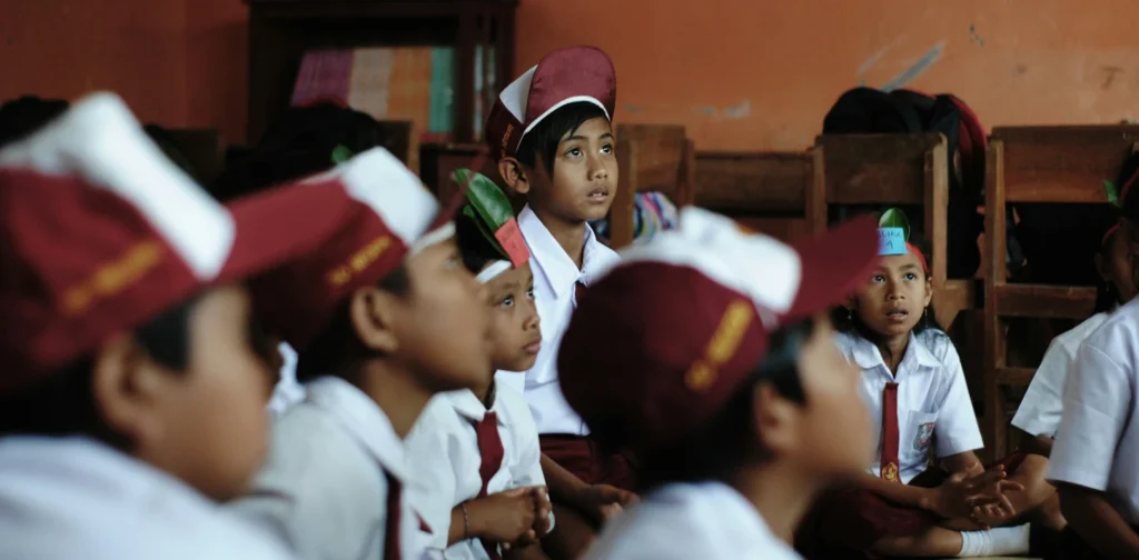 anak-anak sekolah dasar dengan seragam dan topi merah putih duduk di lantai menyimak pelajaran