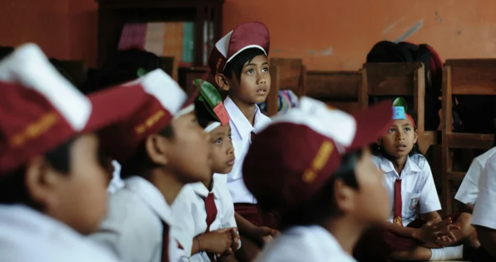 anak-anak sekolah dasar dengan seragam dan topi merah putih duduk di lantai menyimak pelajaran