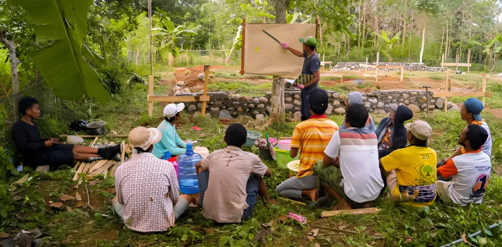 Sekelompok petani duduk beralaskan tanah di ruang terbuka, mengikuti penyampaian materi yang dipandu seorang narasumber yang menggunakan papan tulis.