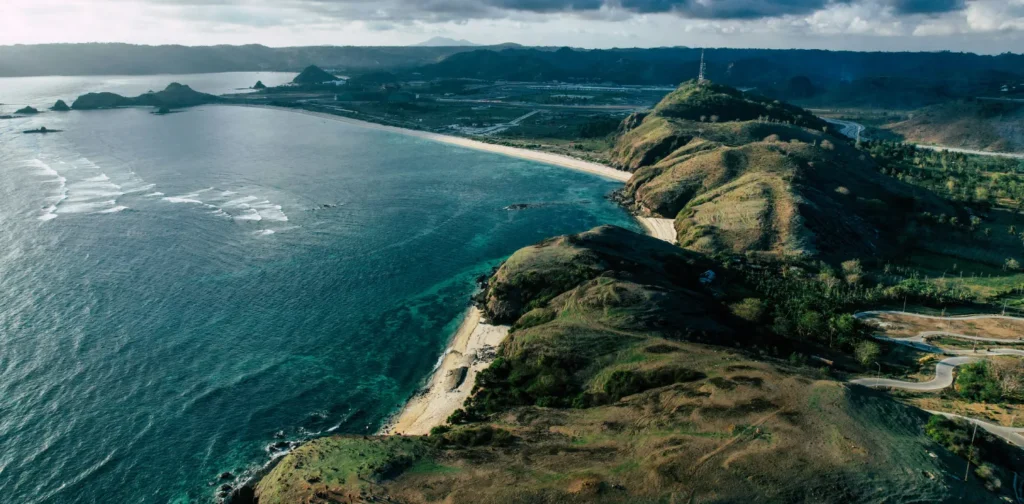 potret pantai dengan air biru laut dan bebukitan hijau dari atas udara