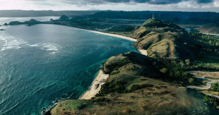 potret pantai dengan air biru laut dan bebukitan hijau dari atas udara