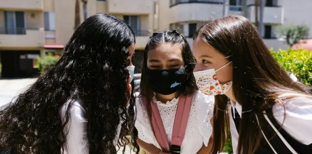 tiga anak perempuan mengenakan masker dengan kepala saling berdekatan seperti sedang berbisik.