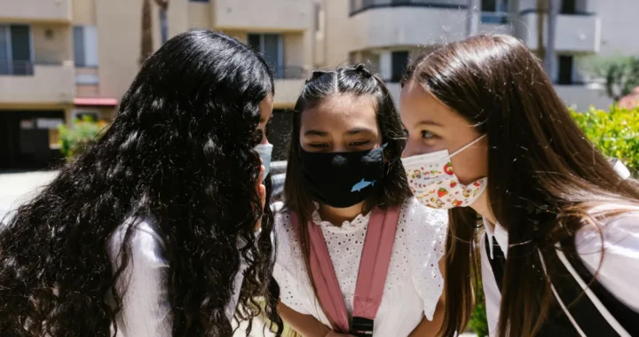 tiga anak perempuan mengenakan masker dengan kepala saling berdekatan seperti sedang berbisik.