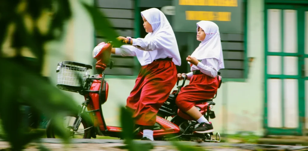 dua anak perempuan berseragam merah putih mengendarai sepeda listrik di jalan.