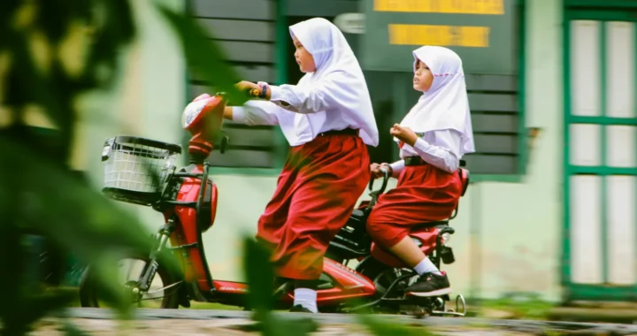 dua anak perempuan berseragam merah putih mengendarai sepeda listrik di jalan.