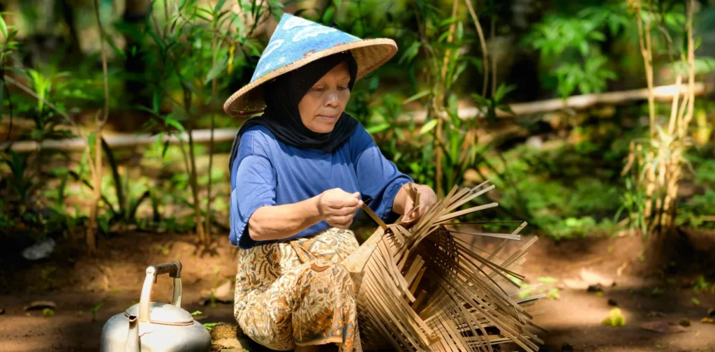 Seorang wanita yang menggunakan topi caping sedang menganyam keranjang bambu.