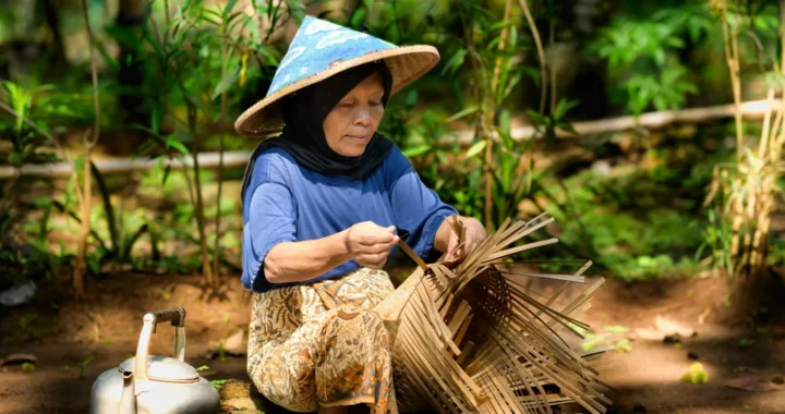 Seorang wanita yang menggunakan topi caping sedang menganyam keranjang bambu.