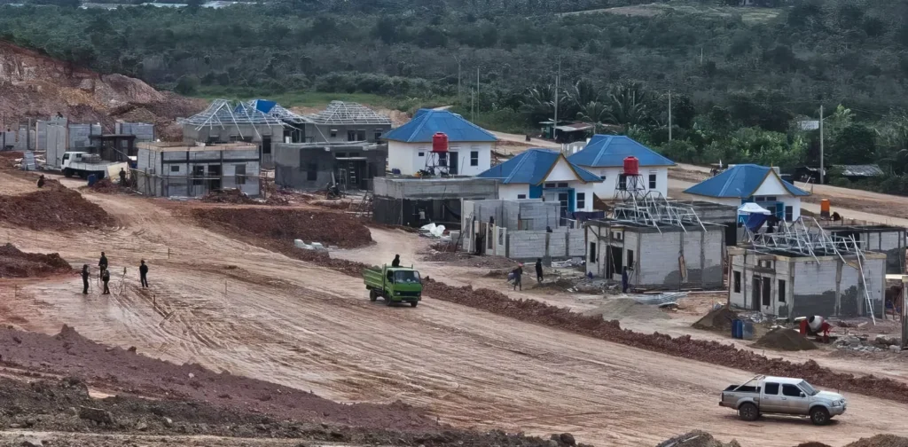 beberapa rumah beratap biru sedang dibangun dengan lalu lalang truk dan orang-orang, latar belakang pepohonan