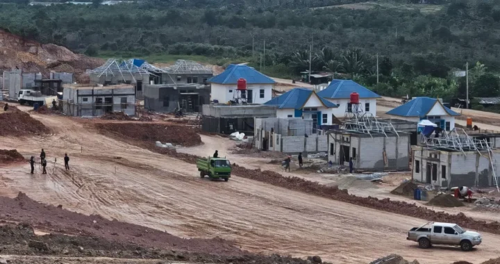 beberapa rumah beratap biru sedang dibangun dengan lalu lalang truk dan orang-orang, latar belakang pepohonan