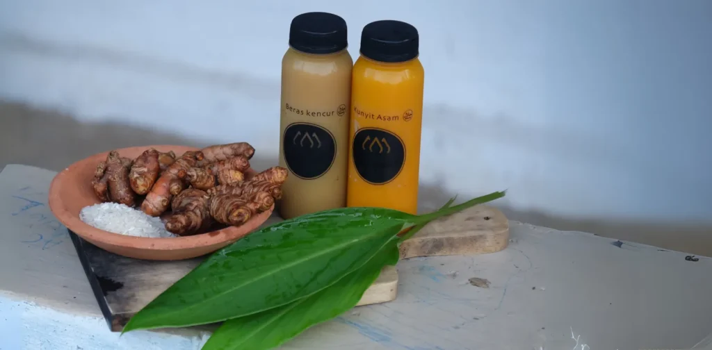 dua botol berisi jamu beras kencur dan kunyit asem serta rempah lainnya di atas talenan kayu