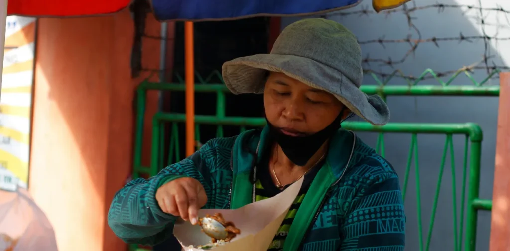 Seorang pedagang sedang memegang pembungkus nasi dan menuangkan lauk makanan.
