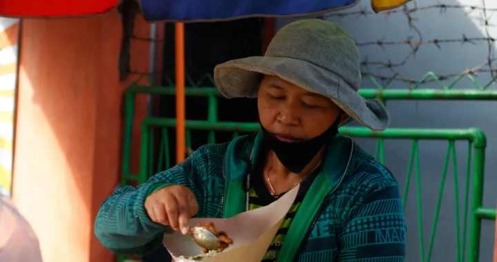 Seorang pedagang sedang memegang pembungkus nasi dan menuangkan lauk makanan.