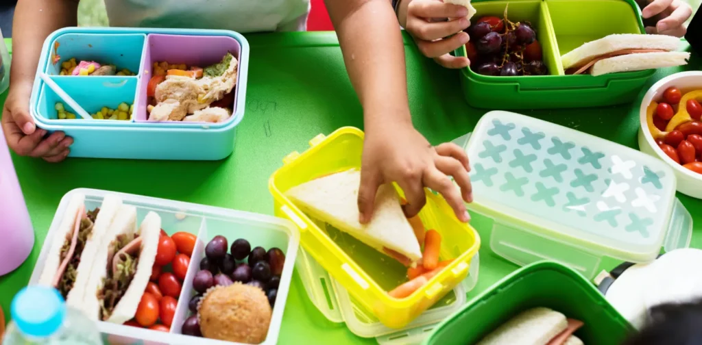 Dua anak sedang memegang roti dengan banyak kotak bekal berisi buah dan berbagai makanan di hadapannya.