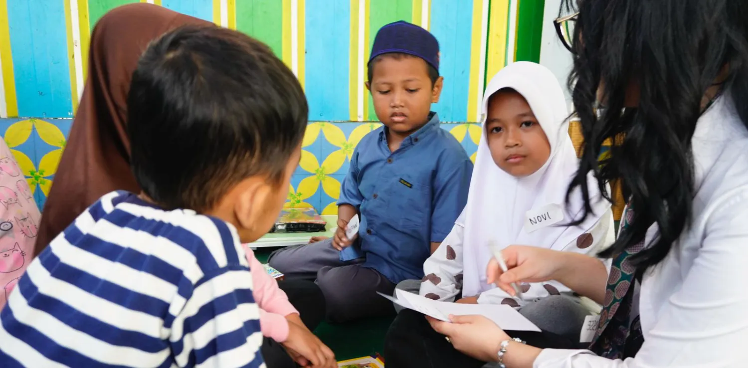 Seorang relawan perempuan bersama empat anak sedang membaca dan menulis bersama dalam lingkaran pada acara literasi Inisiasi Lantara.