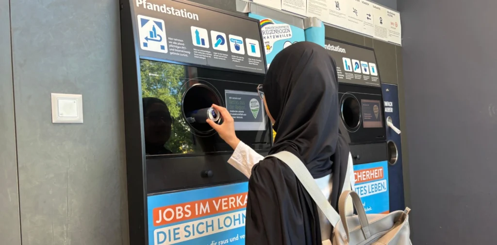 seorang perempuan berjilbab memasukkan kaleng minuman ke dalam mesin daur ulang pfandstation.