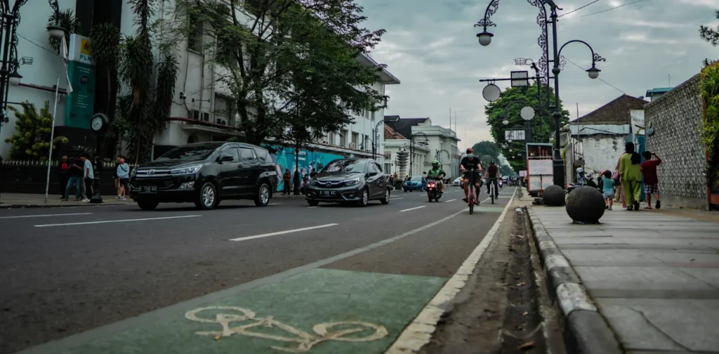 Jalanan dengan mobil, pengendara motor, pengendara sepeda, dan orang-orang yang berjalan kaki di atasnya.