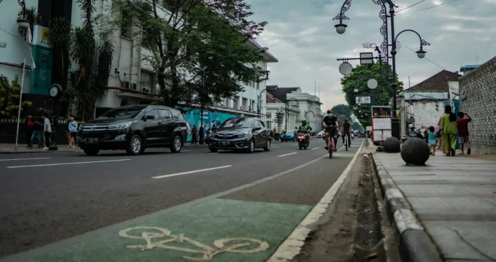 Jalanan dengan mobil, pengendara motor, pengendara sepeda, dan orang-orang yang berjalan kaki di atasnya.