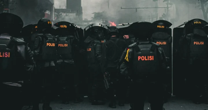 deretan polisi dengan rompi dan tameng menghadap ke arah pengunjuk rasa dengan latar asap hitam