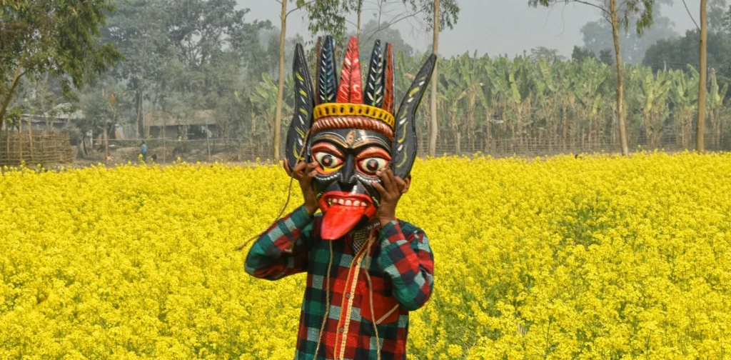 seseorang mengenakan topeng gomira di tengah halaman yang ditumbuhi bunga kuning