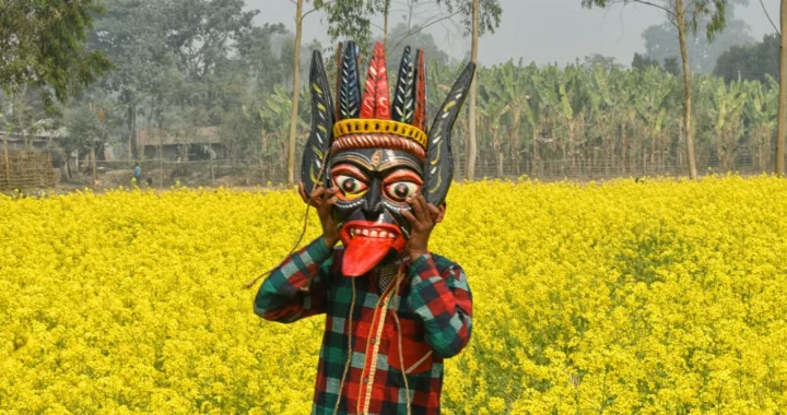 seseorang mengenakan topeng gomira di tengah halaman yang ditumbuhi bunga kuning