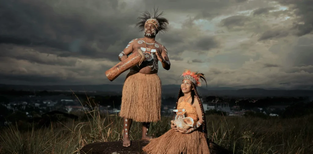 perempuan yang duduk di batang pohon besar, laki-laki berdiri di sampingnya dan dikelilingi rerumputan; keduanya mengenakan pakaian tradisional Papua