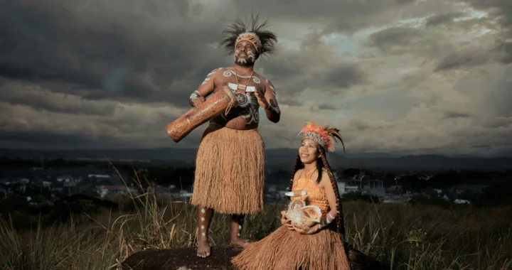 perempuan yang duduk di batang pohon besar, laki-laki berdiri di sampingnya dan dikelilingi rerumputan; keduanya mengenakan pakaian tradisional Papua