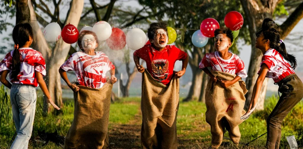 Tiga anak sedang mengikuti lomba balap karung di antara balon yang tergantung, sementara dua anak di samping memberi taburan bedak. Mereka mengenakan kaos merah putih dan berada di jalan tanah di antara pepohonan.