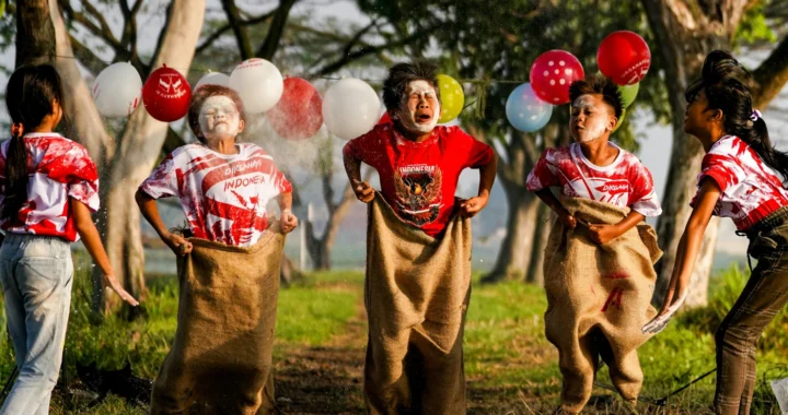 Tiga anak sedang mengikuti lomba balap karung di antara balon yang tergantung, sementara dua anak di samping memberi taburan bedak. Mereka mengenakan kaos merah putih dan berada di jalan tanah di antara pepohonan.