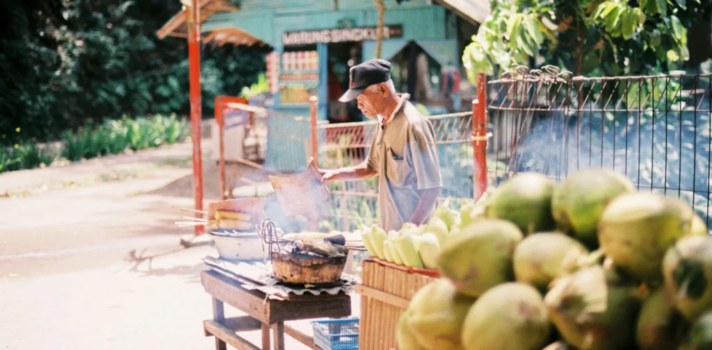 Seorang pria menjual dan mengipas jagung bakar di samping meja yang penuh dengan kelapa muda.