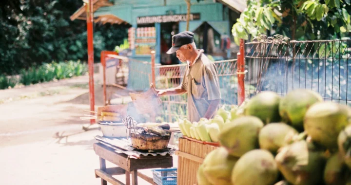 Seorang pria menjual dan mengipas jagung bakar di samping meja yang penuh dengan kelapa muda.