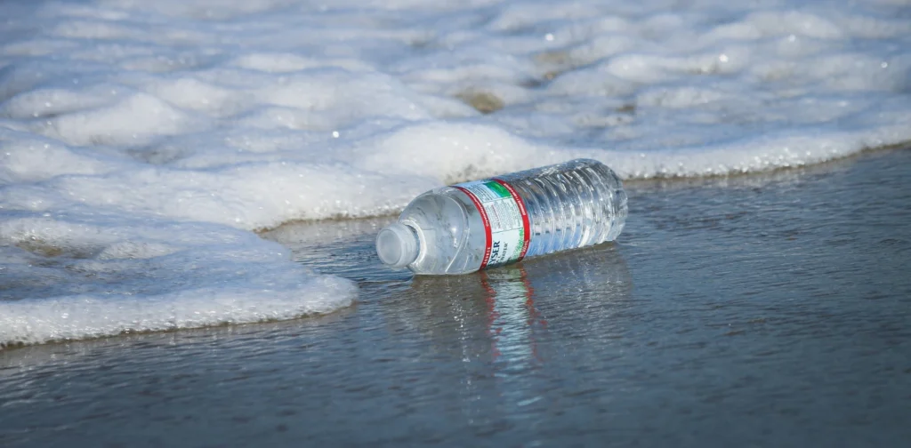 gambar jarak dekat sebuah botol air plastik terdampar di bibir pantai yang berbuih