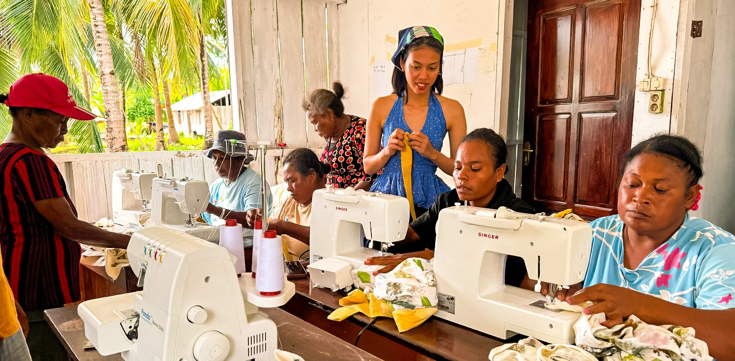 enam perempuan menggunakan mesin jahit, dan satu perempuan berdiri di sisi meja