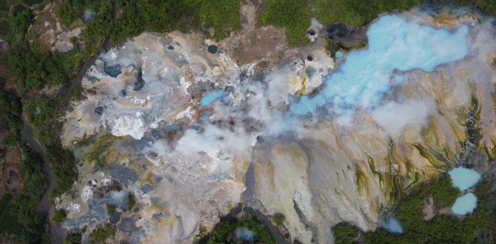 Foto udara salah satu kawasan geothermal di Suoh, Taman Nasional Bukit Barisan Selatan (TNBBS). Menampilkan kawah beruap dengan kolam air berwarna biru dan putih, dikelilingi vegetasi hijau di sekitarnya.