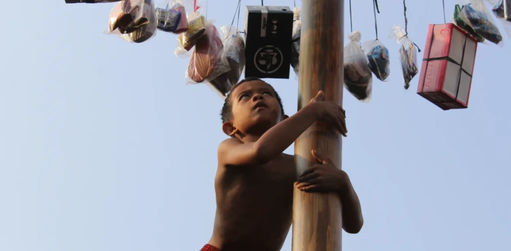 foto seorang anak sedang memanjat batang pinang dan mencoba menggapai hadia-hadiah yang digantung