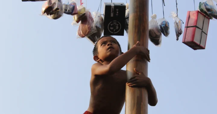 foto seorang anak sedang memanjat batang pinang dan mencoba menggapai hadia-hadiah yang digantung