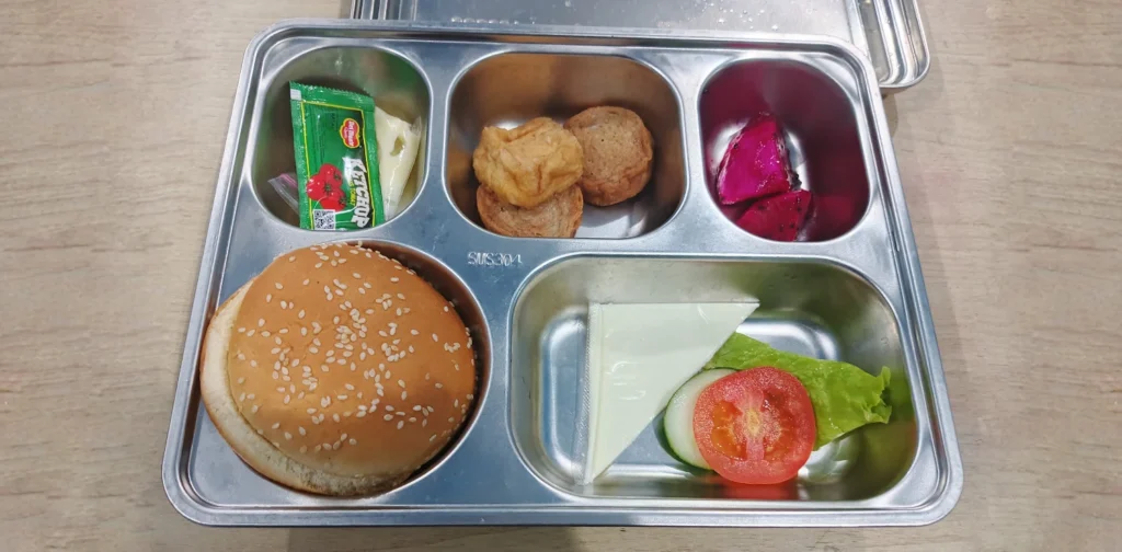 menu makanan dalam wadah kaleng terdiri dari roti, setengah selembar keju, dua potong kecil buah naga, gorengan, dan saus tomat dalam kemasan.