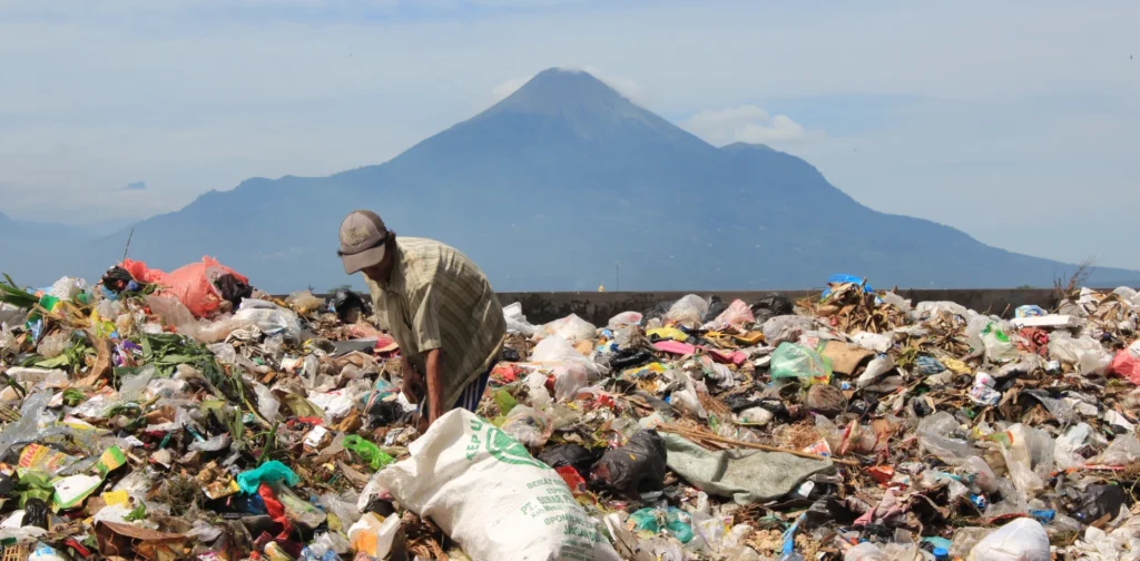 Seorang pria sedang memilah tumpukan sampah di tempat pembuangan terbuka, dengan latar belakang gunung tinggi.